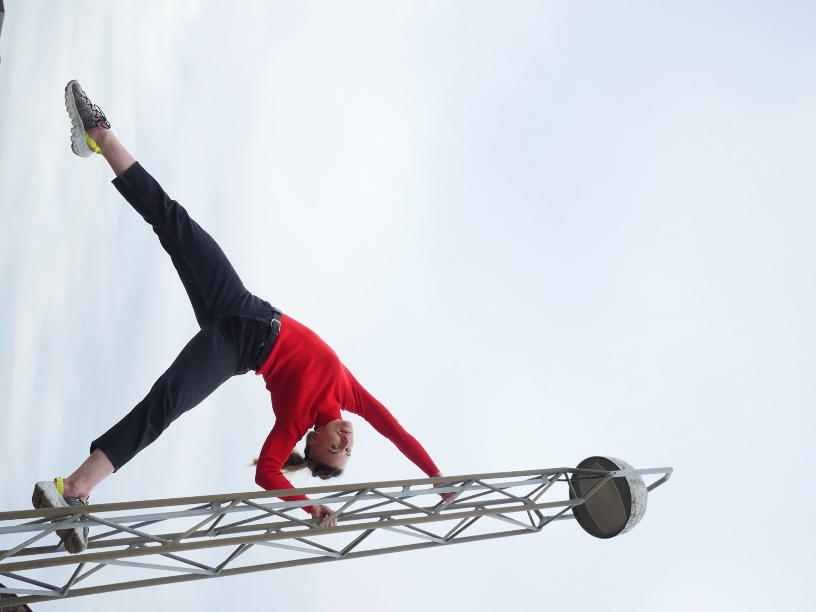 Julia in a wide standing split reaching across to a steel mast against a white sky